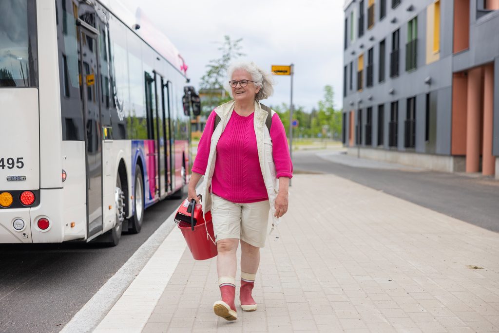 Mummo bussipysäkillä ämpäri ja poimuri kädessä.